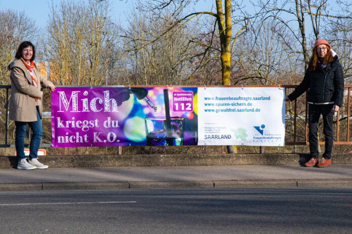 Die Frauenbeauftragte der Stadt Saarlouis, Sigrid Gehl (links), und die Kreisfrauenbeauftragte Astrid Brettnacher an einem der Banner in Saarlouis, die auf die Thematik K.O.-Tropfen hinweisen. Foto: Landkreis Saarlouis/Nicola Schmitt